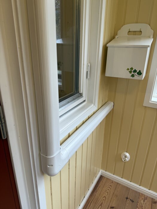 Målat innertak med linoljefärg, vitmålad kabelkanal och gammal lampett bredvid ett fönster. Gult träpanel på väggen.