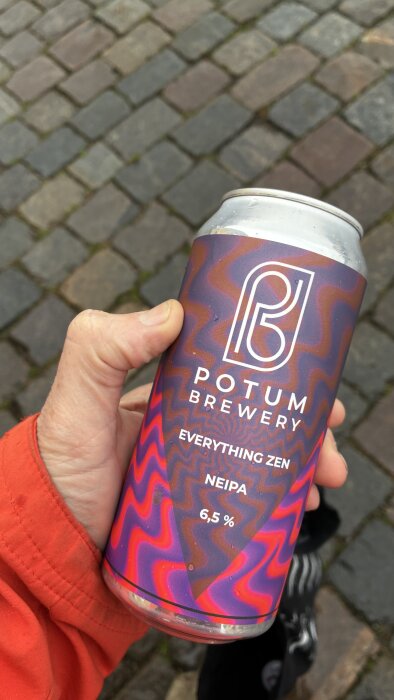 Hand som håller en färgglad burk med öl från Potum Brewery med namnet "Everything Zen NEIPA" och alkoholhalten 6,5 %, bakgrund av stenläggning.