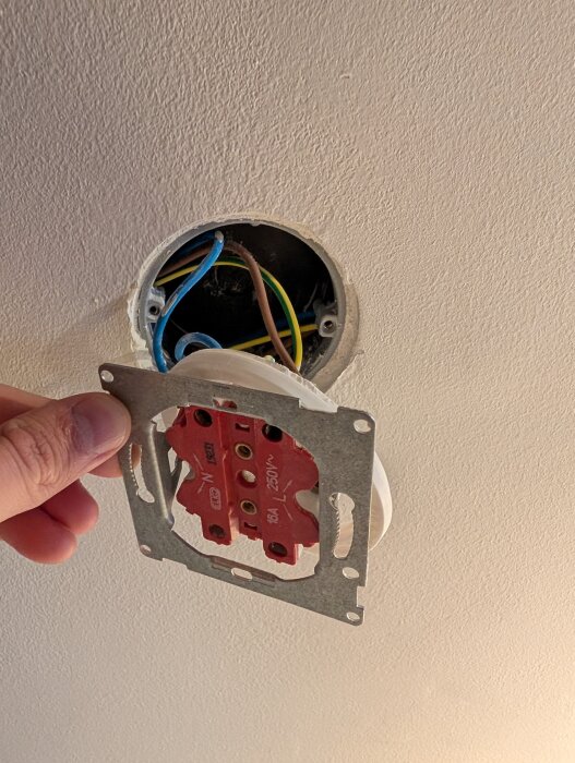 Hand installerar en eluttag bakom en vägg med synliga kablar i olika färger i ett rör inuti väggen.