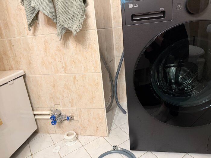 Tvättmaskin bredvid vägg med tappventil och rör. Grå slangar intill tvättmaskinen och en tejprulle på golvet.