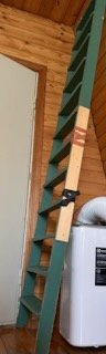 Grön trappa till loft i ett rum med trätak och vit vägg; synlig del av vit hushållsapparat intill trappan.