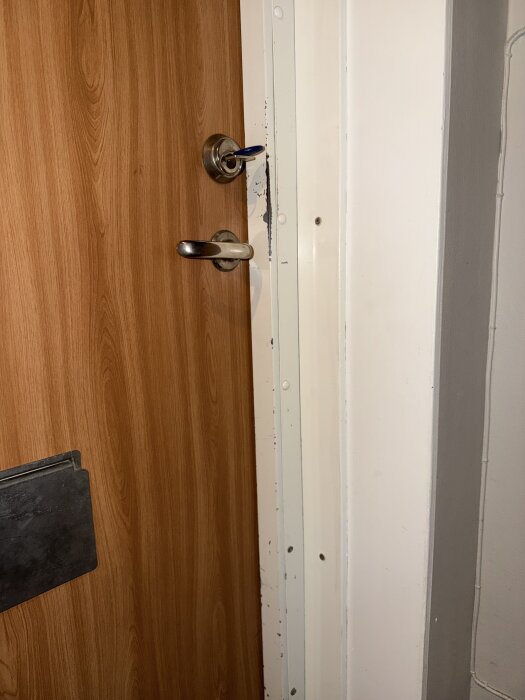 Lägenhetsdörr med träfinish och dörrkarm som visar glipa mellan dörr och karm. Dörrbladet har handtag och nyckel i låset.