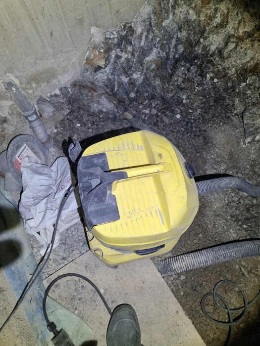 Gul industridammsugare på byggarbetsplats, omgivet av stenig mark och betong, med ventilation och kabel på golvet.