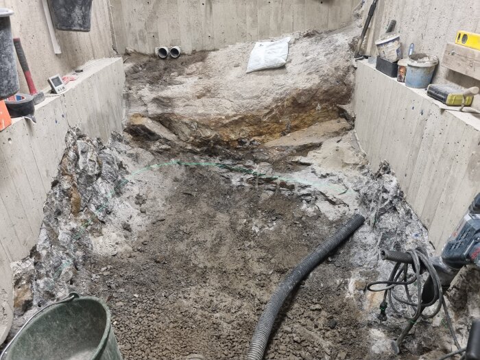 Byggarbetsplats i en grund med betongväggar, synlig berggrund och verktyg som borrmaskin och rör. Sand och grus på marken.