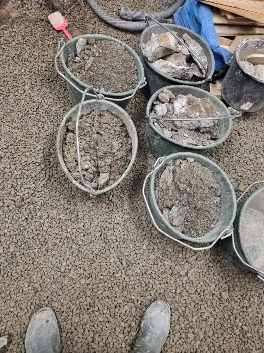 Sex hinkar med grus och stenblock på en grusbelagd yta, bredvid en röd spade och byggmaterial i bakgrunden.