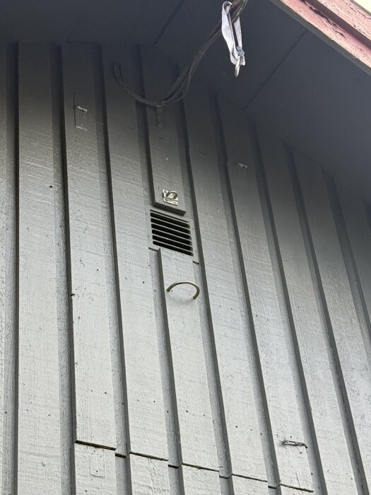 Yttre fasad med horisontell ventilgaller och krok under. Högre upp hänger ett tvinnat rep eller rem från takutsprånget.