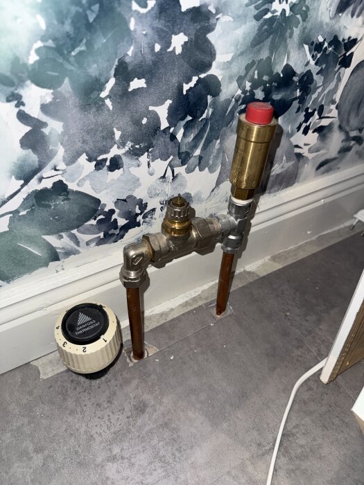 Radiatorventil från Danfoss med numrerad termostat före en blommig tapet. Kopparrör leder till ventilen som sticker ut från golvet.