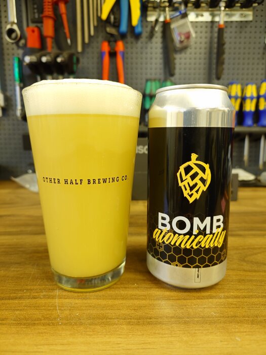 Ett glas ljus öl från Other Half Brewing Co. bredvid en ölburk med texten "Bomb Atomically," stående på en träyta med verktyg i bakgrunden.