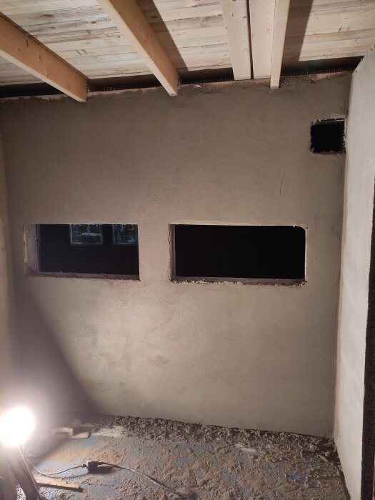 Nypustad vägg med två rektangulära fönsteröppningar och träbjälkar i taket, lampa lyser upp arbetsområdet.