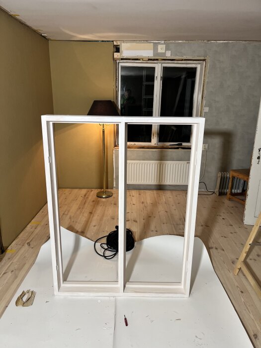 Målat fönster utan glas på golvet, med gul vägg, fönster och lampa i bakgrunden, inomhusmiljö under renovering.