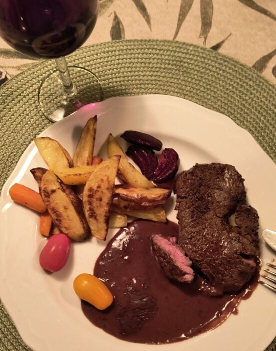 En tallrik med kött, stekta potatisklyftor, färgglada grönsaker och mörk sås. Ett glas rödvin står bredvid på en grön bordsduk.