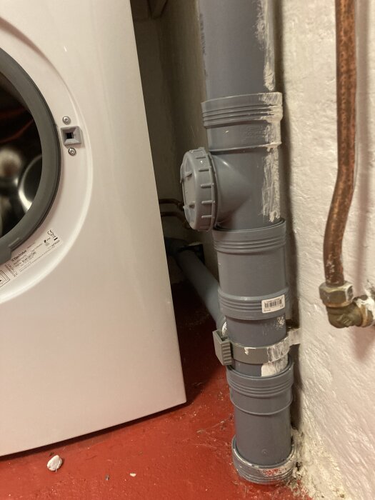 Grått avloppsrör och kopparrör intill en vit tvättmaskin på ett rött betonggolv i en tvättstuga.