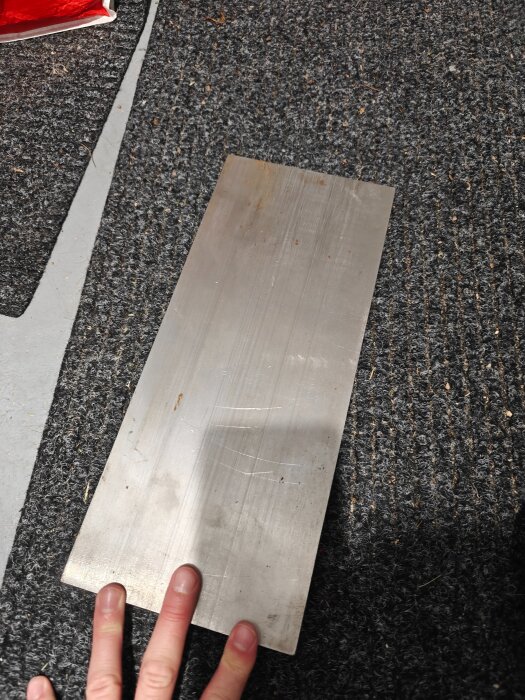 En rektangulär stålbit på en grå matta, delvis stödjad av en hand.