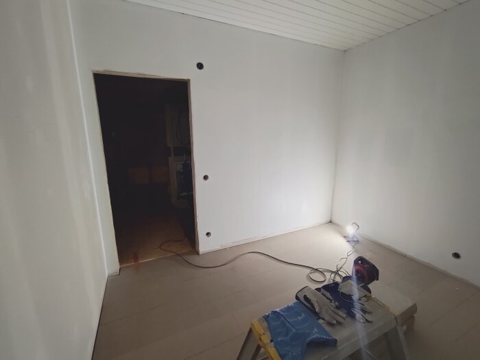 Tillbyggnad invändigt med nybyggda och målade väggar, stark arbetslampa lyser, verktyg och handskar ligger på en stege.