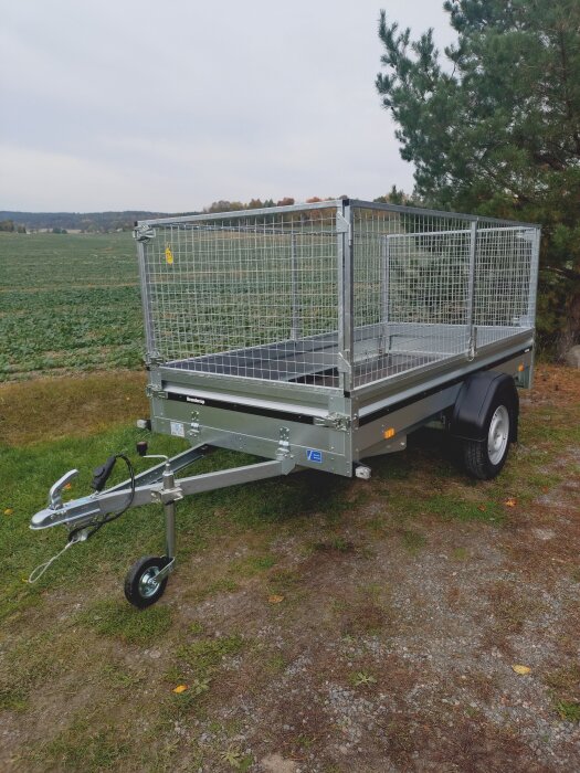 En obromsad släpvagn med galler är parkerad på en grusplan vid en åker, omgiven av träd.