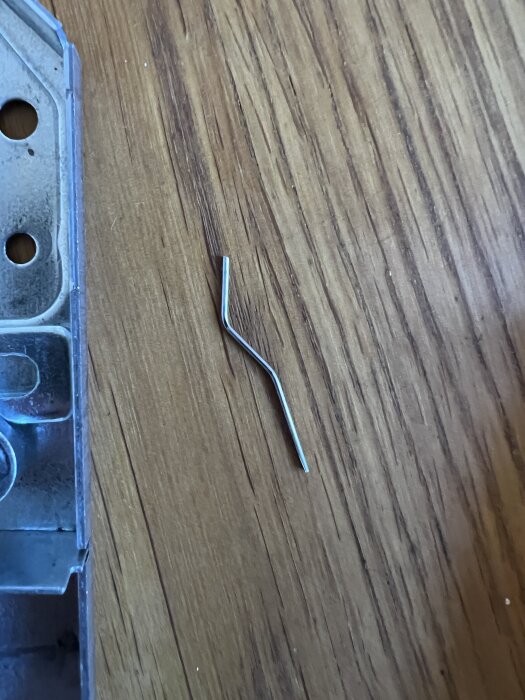 Liten böjd metallbit på ett bord bredvid en del av ett lås, troligen från en ASSA 8765.