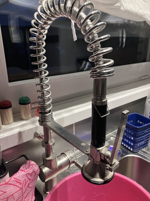 Köksspruta i rostfritt stål ovanför en rosa hink, med diskbänk i bakgrunden.
