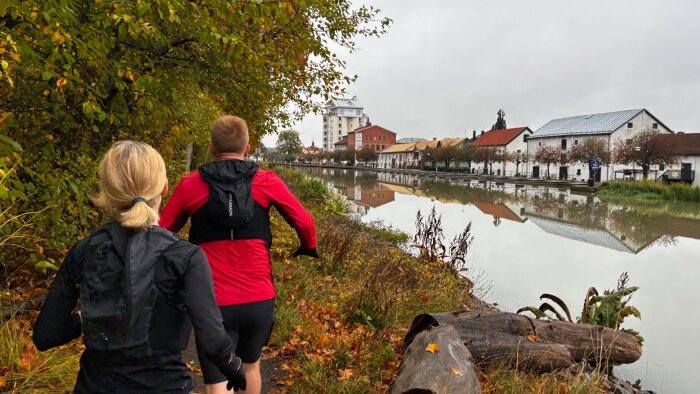 Två personer joggar på en stig bredvid en kanal med spegelblanka vattenytor och byggnader i bakgrunden, omgivna av höstfärgade träd.