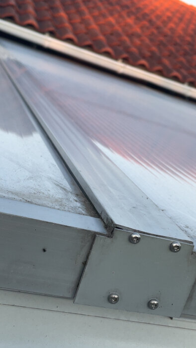 Täckbit med flera skruvar i aluminiumprofil på ett kanalplasttak mot tegelpannor, med fokus på monteringsdetalj.