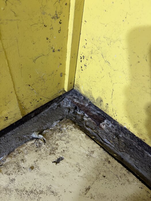 Gul vägg med sprickor och vittrad grund i ett hörn av ett förråd från 60-talet; synliga mindre mellanrum och en snigel i hörnet.