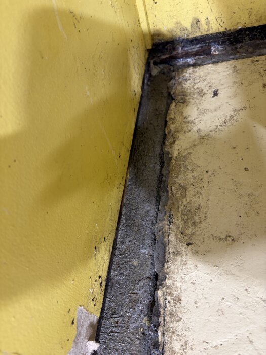 Spricka mellan betonggolv och vägg i ett äldre förråd med gulmålad vägg. Grunden verkar vittrad och det finns utrymme mellan strukturerna.