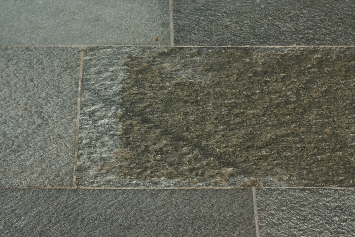 Mörkgrå stengolvplattor med olika texturer, placerade kant i kant. En del av plattorna har en blöt yta som ger en mörkare nyans.