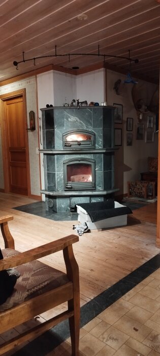 Braskamin i ett rum med träpanel, stenarbeten och oljad trädörr, med eld synlig i kaminens lucka. En låda med filt ligger vid sidan.