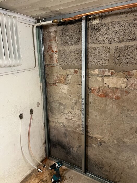 Renovering av källarvägg från 1932 med stålreglar, synligt tegel och betong. Rör och kabel synliga, samt två anslutna vattenslangar och en borrmaskin på golvet.