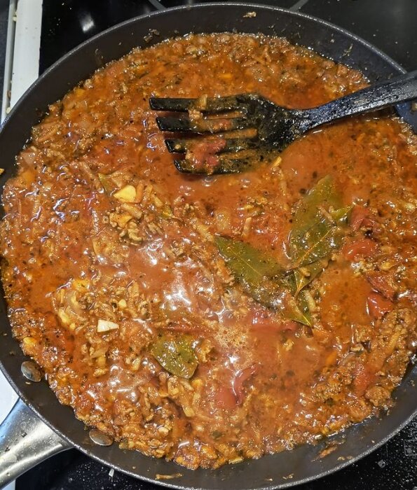 Köttfärssås som kokar i en stekpanna med tomater, kryddor och lagerblad, en svart stekspade ligger ovanpå såsen.