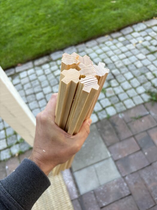 Hand håller träbitar i form av spröjs till innerbågar, stående på stenlagd yta med gräsmatta i bakgrunden.