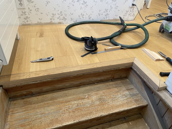 Träslutning vid trappa med verktyg som slipmaskin och hobbykniv på parkettgolv.