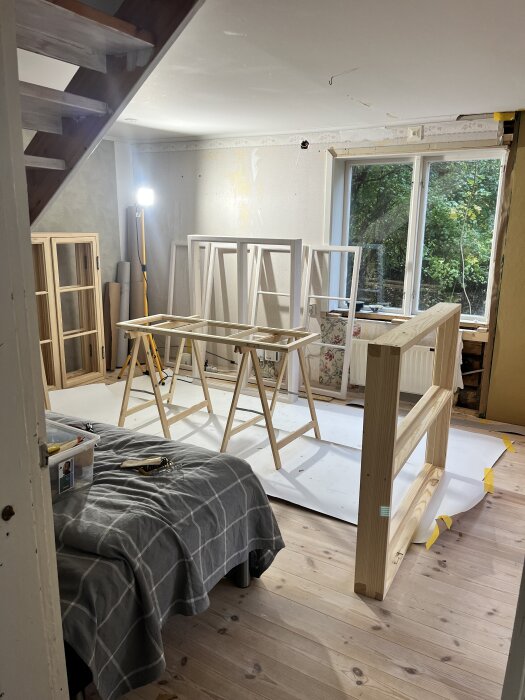 En renoveringsplats inomhus med fönsterkarmar på bockar, ett täckt bord och en lampa står i ett rum med fönster mot en trädgård.