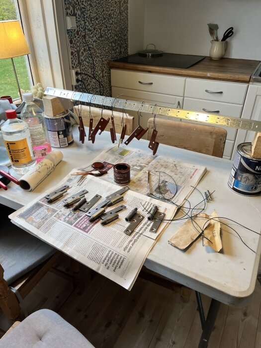 Gångjärn målade med järnmönja hänger på tork över ett bord täckt med tidningar, burkar med färg, verktyg och flaskor i ett kök.