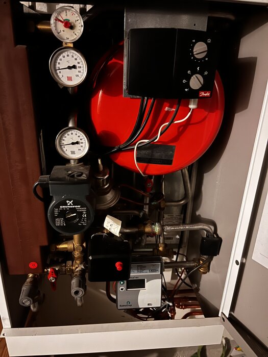 Fjärrvärmecentral med tryckmätare, röda kärl och termostat, justerad för bättre uppvärmning då elementen förblir kalla i huset trots låg utetemperatur.