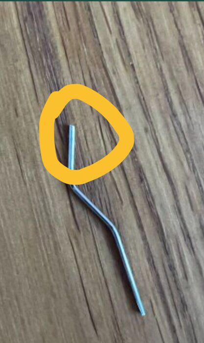 Böjd metalltråd ligger på ett trägolv, med en del av tråden markerad med en gul cirkel.