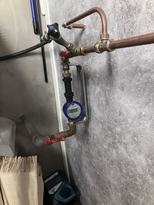 Vattenmätare monterad på vägg med kopparrör och ventiler i ett installationsrum.