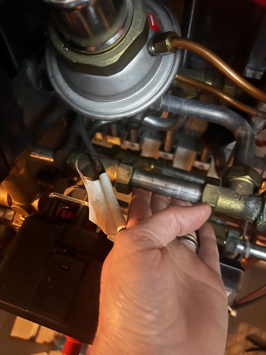Hand som håller ett metallrör under en teknisk installation, med ett svart fyrkantigt objekt synligt nedanför.