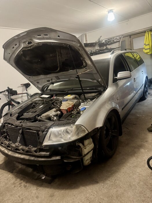 Silverfärgad bil med öppen motorhuv i garage, cykel står bredvid och en varselväst hänger på väggen.
