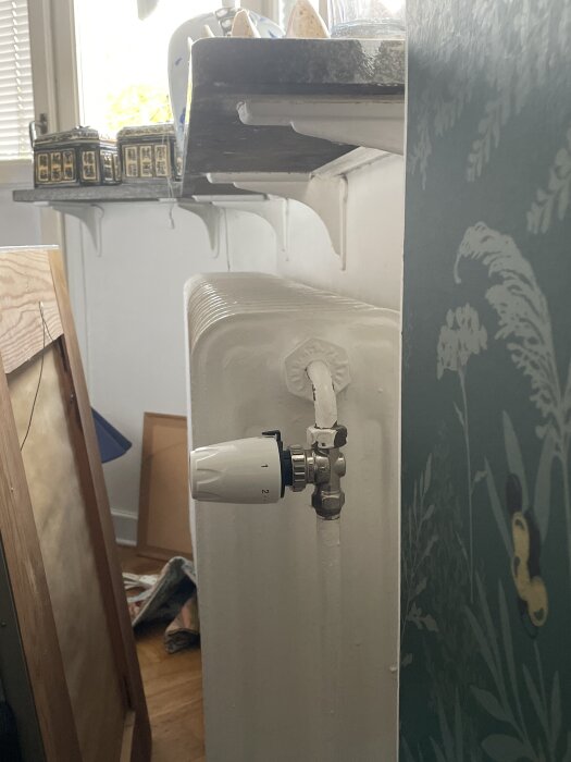 Radiator vid burspråk, sticker ut från fönsterkarmen, termostat synlig. Elementet är vitmålat, omgiven av grön tapet med växtmotiv.