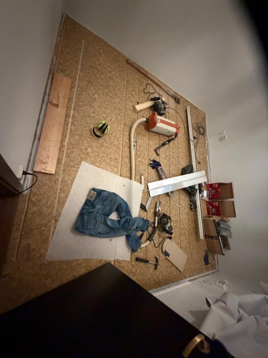 Verktyg och material för renovering spridda på ett golv, inklusive en orange maskin, skyddshjälm, jeans, en såg, och andra småverktyg.