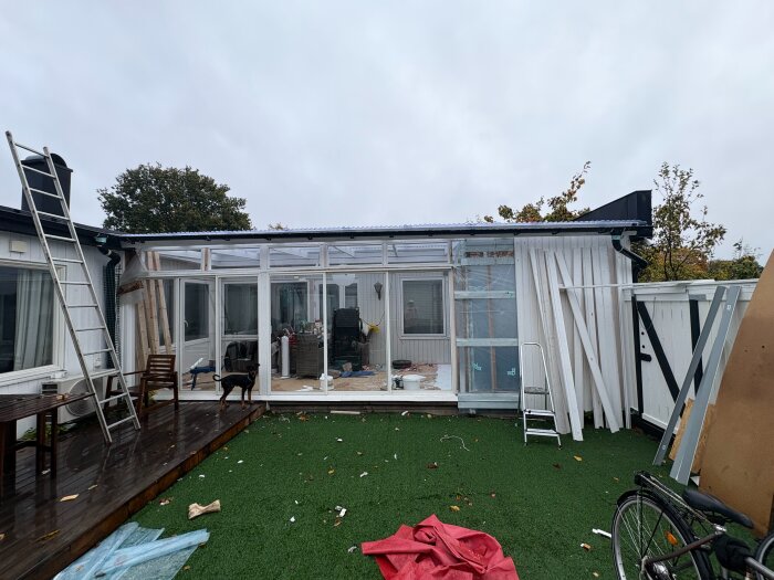 Radhusets altan med genomskinligt tak, stolar, möjlig pågående renovering och en hund på konstgräsmatta. Stege mot väggen.