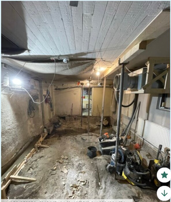 Renovering av källarrum med tomt betonggolv, exponering av rör och elkablar. Arbetslampor lyser upp rummet, olika verktyg ligger utspridda.