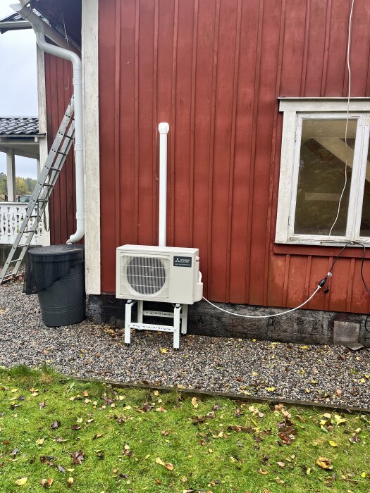 Luftvärmepump av märket Mitsubishi Ninja installerad på röd husfasad, omgiven av grus och grönska, med sladd provisoriskt ansluten.