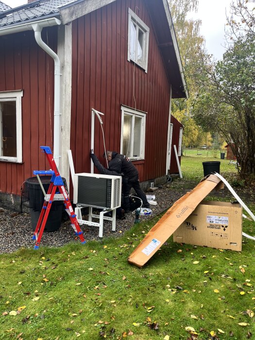 Installation av en luftvärmepump utomhus på ett rött hus, med en person som arbetar och utrustning runtomkring på marken.