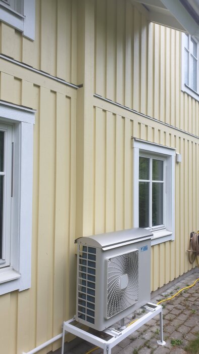 Gul husfasad med fönster, luftvärmepump installerad på en vit ram, delvis dold av panelbrädor för att täcka plastkanal.