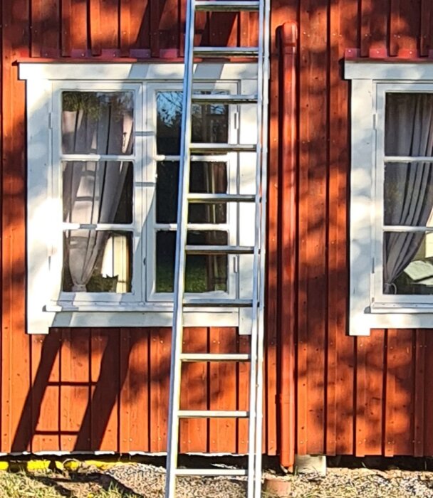 Stege placerad mot fasaden av ett rött trähus med vita fönster och gardiner.