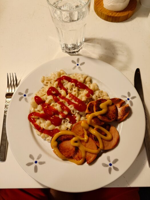 Tallrik med makaroner med ketchup och stekt korv med senap, samt ett glas vatten bredvid.