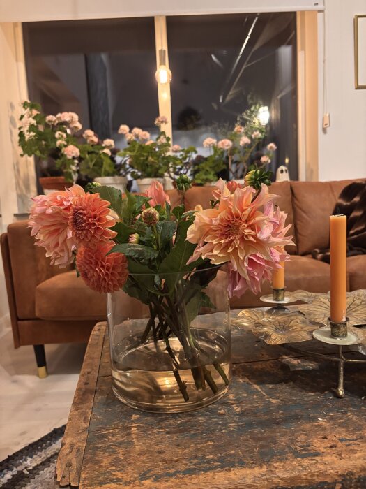 Vas med dahlior och pelargoner på ett rustikt träbord i ett vardagsrum med soffa och fönster i bakgrunden.
