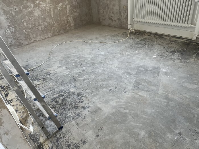 Betonggolv i ett rum med avskalad yta, spår av gammalt lim syns. En stege står lutad mot väggen och ett element i bakgrunden.