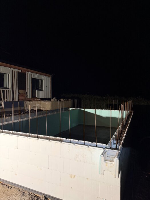 En byggplats under kvällstid där armeringsjärn sticker upp från block runt en blivande pool som är belyst av bygglampor.